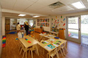 A photo shows a daycare table on hardwood floors with teal, orange and green shaped toys on top. People stand in the distance chatting.