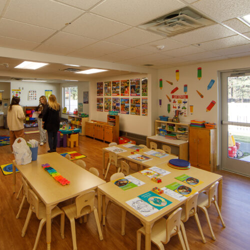 A photo shows a daycare table on hardwood floors with teal, orange and green shaped toys on top. People stand in the distance chatting.