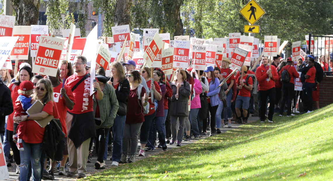 People wearing red shirts and holding signs that read "Tacoma on strike" walk beside a grassy area outdoors.