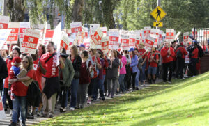 People wearing red shirts and holding signs that read "Tacoma on strike" walk beside a grassy area outdoors.