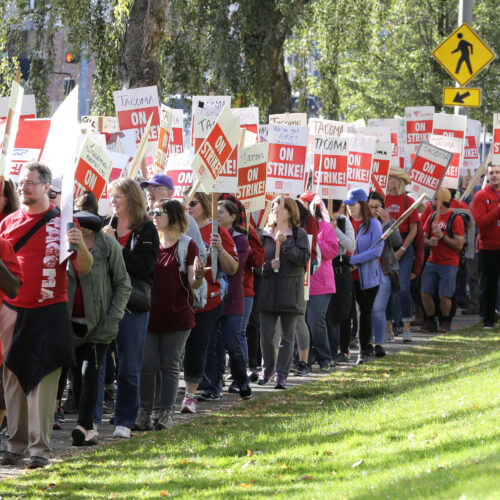 People wearing red shirts and holding signs that read "Tacoma on strike" walk beside a grassy area outdoors.