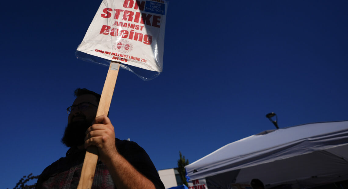 A Boeing worker waves a picket sign. The sign reads, "on strike against Boeing." There is blue sky in the background.