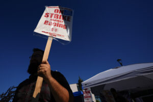 A Boeing worker waves a picket sign. The sign reads, "on strike against Boeing." There is blue sky in the background.