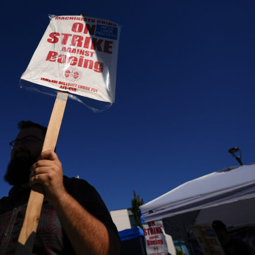 A Boeing worker waves a picket sign. The sign reads, "on strike against Boeing." There is blue sky in the background.