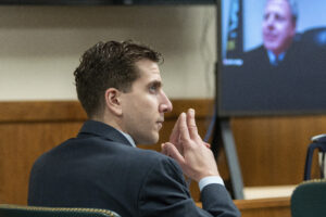 A side profile of Bryan Kohberger in a suit shows him sitting in a courtroom with his fingers pressed together.