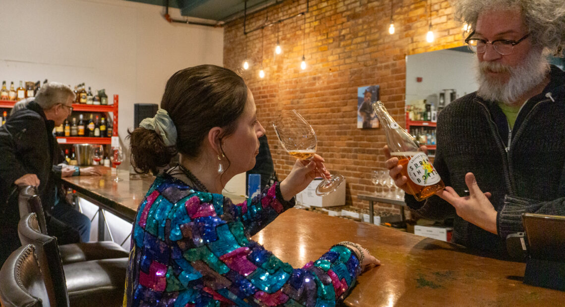 A woman in a sparkly jacket sits at a wooden bar and sips a glass of pink wine.