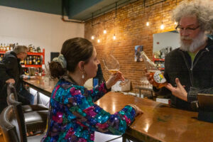 A woman in a sparkly jacket sits at a wooden bar and sips a glass of pink wine.