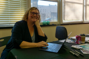 A blonde woman in glasses sits at her desk with a laptop