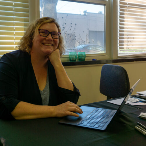 A blonde woman in glasses sits at her desk with a laptop