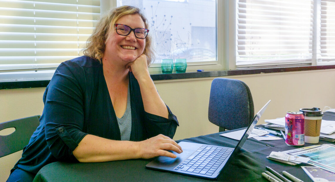 A blonde woman in glasses sits at her desk with a laptop.