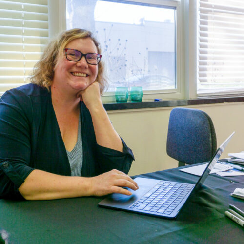 A blonde woman in glasses sits at her desk with a laptop.