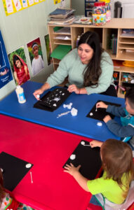 A woman with dark hair and a blue shirt sits at a blue and red table with young children working on a craft project together.