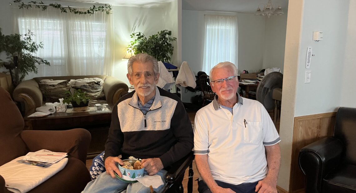 Two men are seated inside a house. Several armchairs are seen in the background.