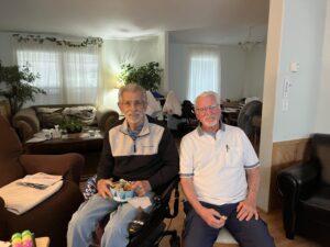 Two men are seated inside a house. Several armchairs are seen in the background.