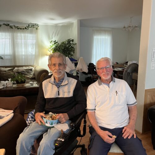 Two men are seated inside a house. Several armchairs are seen in the background.