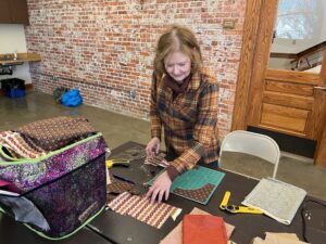 A person in an orange and black plaid jacket looks at fabric laid out across a table. Behind them, there is a brick wall and wooden door.