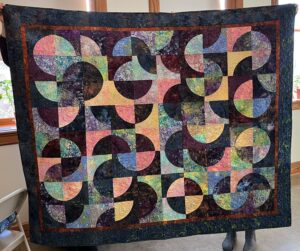 A finished quilt is held up. The quilt has a black background and multi-colored swirls in its design.