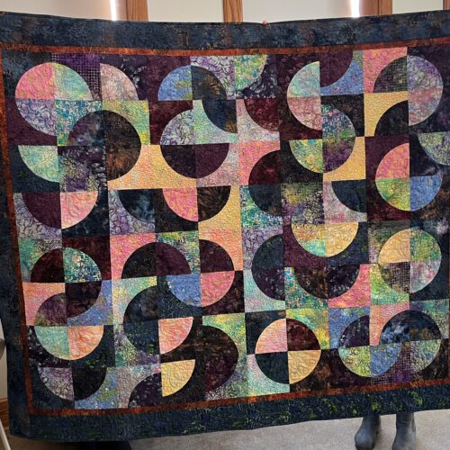 A finished quilt is held up. The quilt has a black background and multi-colored swirls in its design.