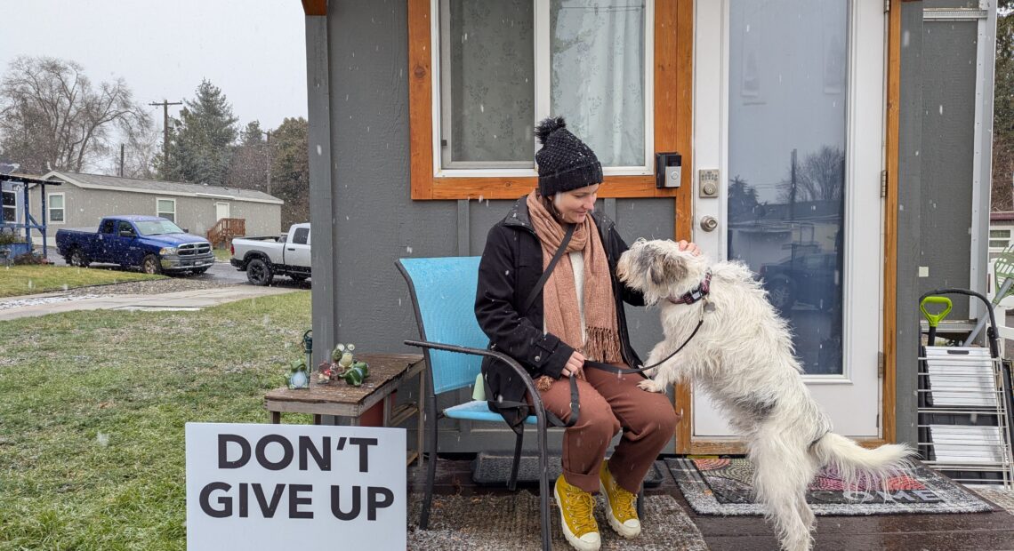 A person with orange pants, a black jacket and black hat sits in a chair in front of their grey tiny house with a white dog jumping on their lap. A sign next to them reads "Don't Give Up."