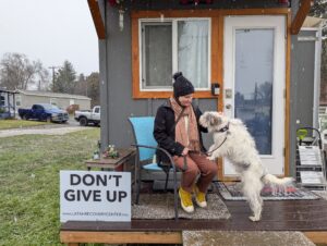 A person with orange pants, a black jacket and black hat sits in a chair in front of their grey tiny house with a white dog jumping on their lap. A sign next to them reads "Don't Give Up."