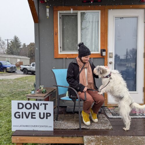 A person with orange pants, a black jacket and black hat sits in a chair in front of their grey tiny house with a white dog jumping on their lap. A sign next to them reads "Don't Give Up."