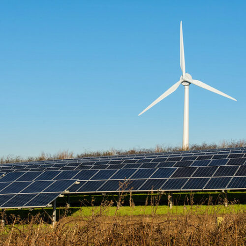 A field of solar panels is in the bottom third of the picture. The solar panels sit on top of green-ish, brown-ish grass. There is a single, white wind turbine behind the solar panels. The sky is a light blue.