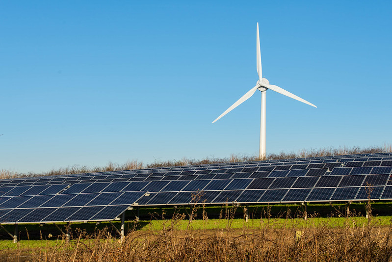 A field of solar panels is in the bottom third of the picture. The solar panels sit on top of green-ish, brown-ish grass. There is a single, white wind turbine behind the solar panels. The sky is a light blue.