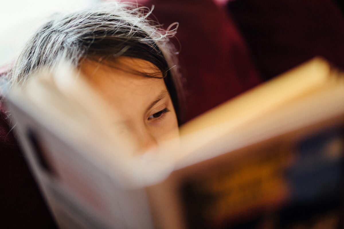 A young child with brown hair holds open a book in a way that obscures most of their face.