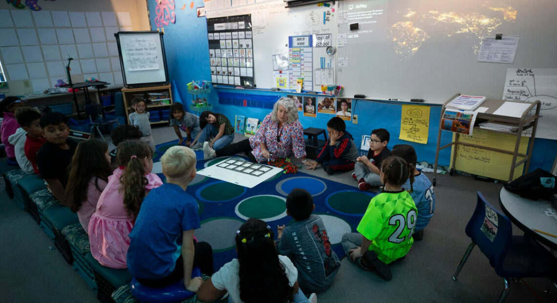 A woman sits on the ground in the front of a classroom. A group of children are gathered around her.