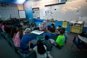 A woman sits on the ground in the front of a classroom. A group of children are gathered around her.