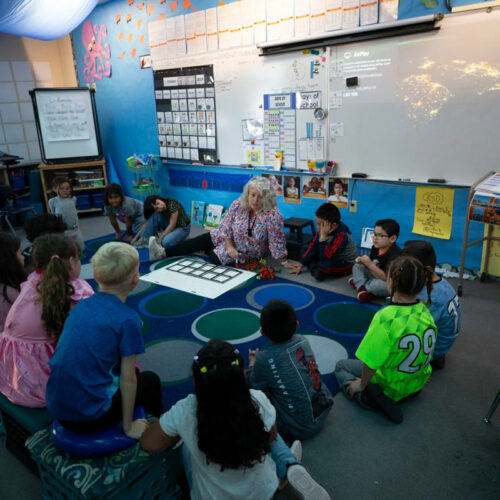 A woman sits on the ground in the front of a classroom. A group of children are gathered around her.