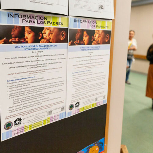 Flyers in English and Spanish are viewed up close. They are posted on a wall. In the background, a person walks by.