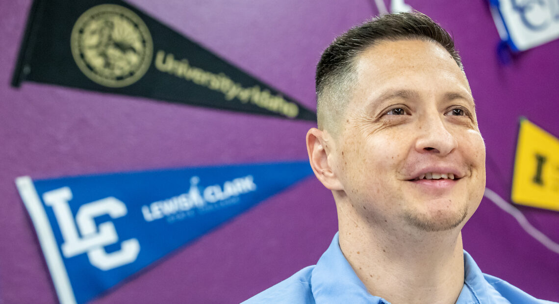 A man in a blue shirt stands in front of a dark purple wall. The wall has college pennants displayed on it.