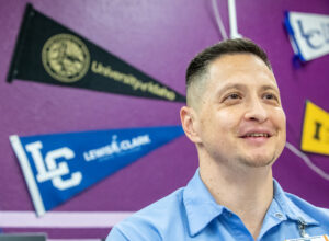 A man in a blue shirt stands in front of a dark purple wall. The wall has college pennants displayed on it.