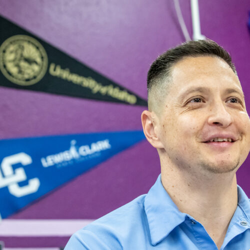 A man in a blue shirt stands in front of a dark purple wall. The wall has college pennants displayed on it.