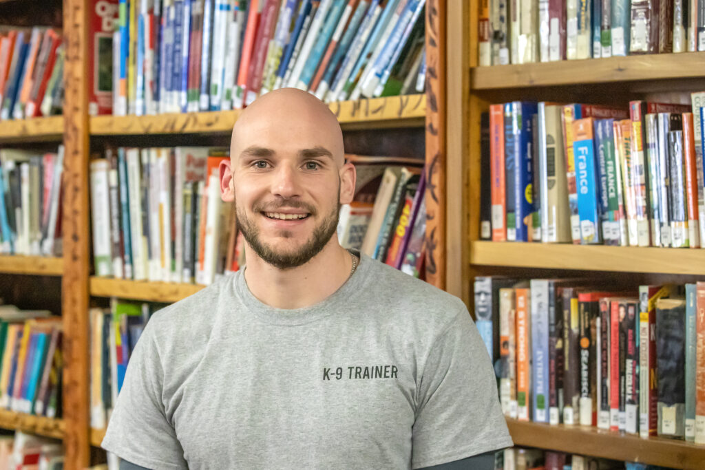 A man in a gray shirt stands in front of a series of wooden bookcases. They hold an assortment of books.