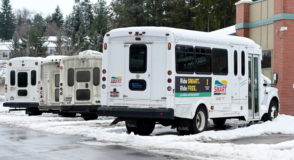 A row of small, white buses are parked outside in a parking lot. There is snow on the ground.