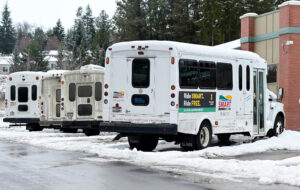 A row of small, white buses are parked outside in a parking lot. There is snow on the ground.