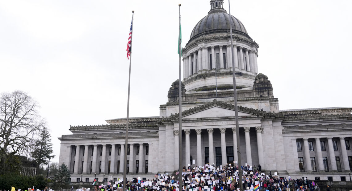 A large group of people, some holding signs, stand in front of the state capitol building in Olympia, Washington.