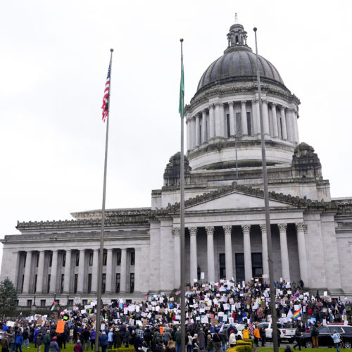 A large group of people, some holding signs, stand in front of the state capitol building in Olympia, Washington.