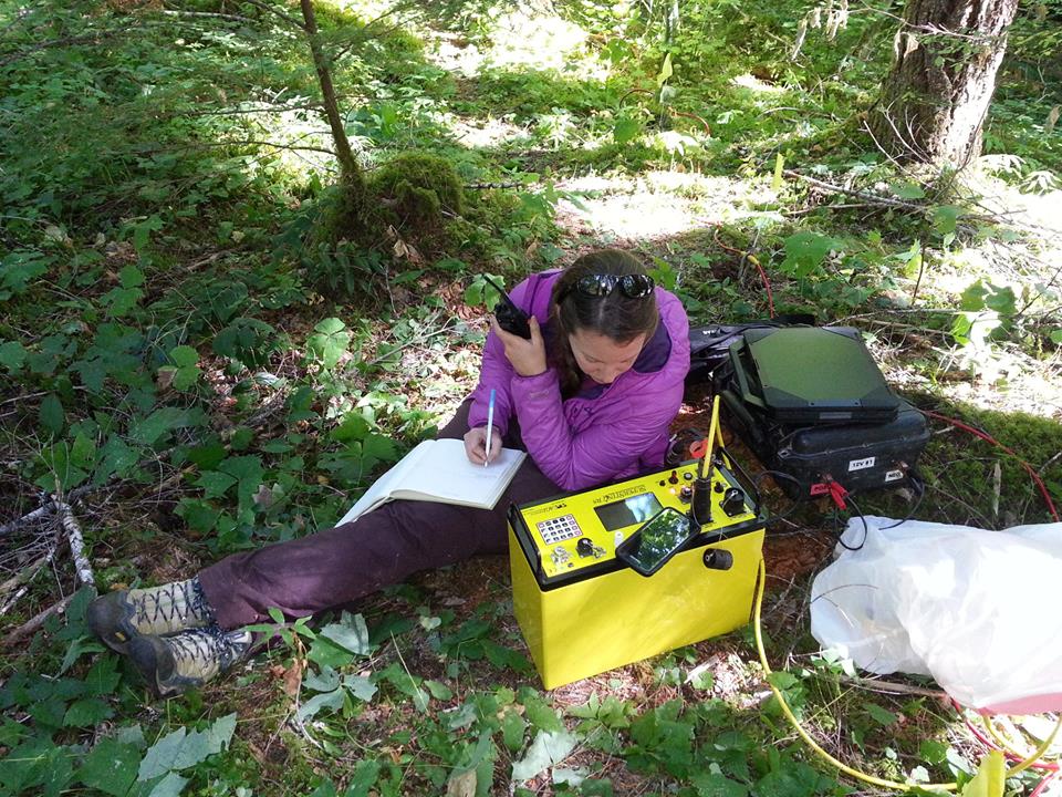 A woman with a purple shirt and purple pants monitors activity on a yellow scientific device while sitting on the ground surrounded by mountain greenery.