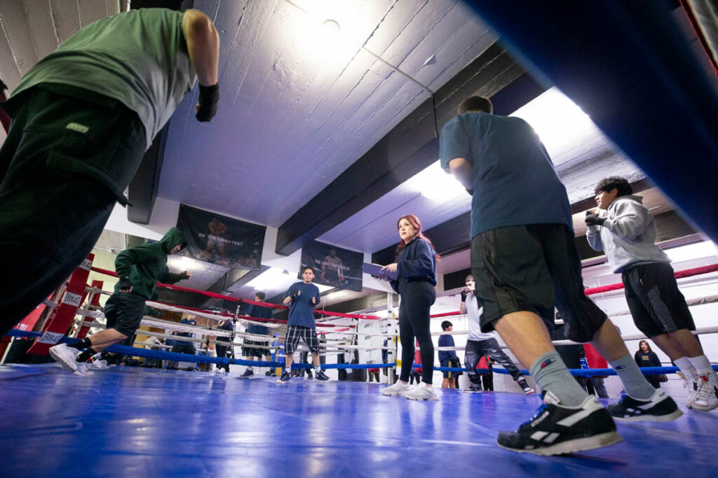 A woman in a blue sweatshirt and black leggings holds a clipboard. She is standing in the middle of a blue boxing ring. A group of people stand around her.