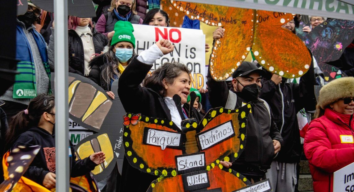 A person holds a sign shaped like an orange butterfly. The words "parque padrinos" and "stand with healthcare, immigrants, unemployment" are written on the sign. A crowd stands behind them.