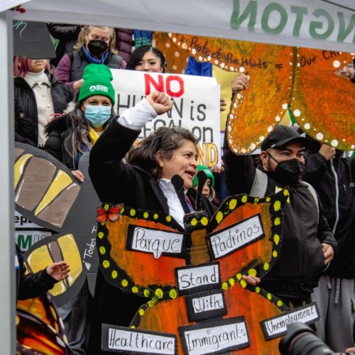 A person holds a sign shaped like an orange butterfly. The words "parque padrinos" and "stand with healthcare, immigrants, unemployment" are written on the sign. A crowd stands behind them.