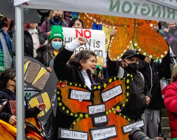 A person holds a sign shaped like an orange butterfly. The words "parque padrinos" and "stand with healthcare, immigrants, unemployment" are written on the sign. A crowd stands behind them.