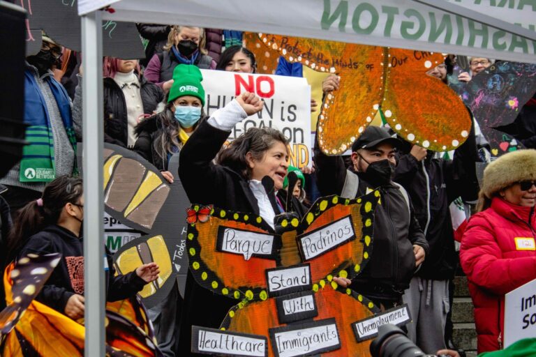 A person holds a sign shaped like an orange butterfly. The words "parque padrinos" and "stand with healthcare, immigrants, unemployment" are written on the sign. A crowd stands behind them.