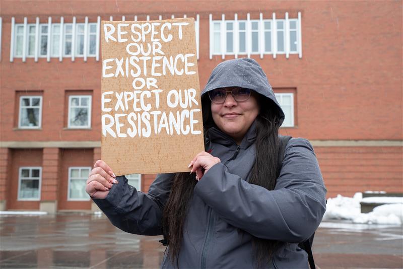 A person holds up a cardboard sign that reads: " Respect our existence or expect our resistance."