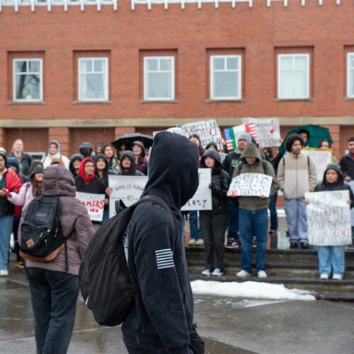 A person in a black sweatshirt walks by a group of people protesting. The protesters are holding signs.