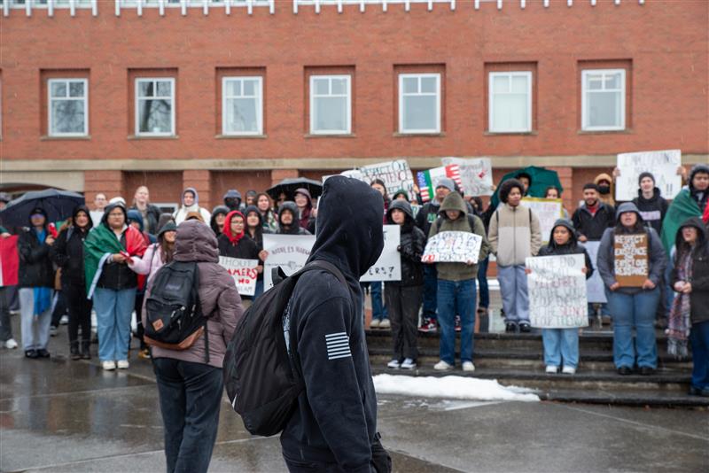 A person in a black sweatshirt walks by a group of people protesting. The protesters are holding signs.