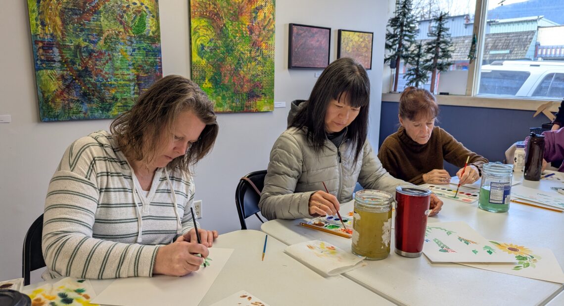 Three women sit at a white table painting flowers on watercolor paper.